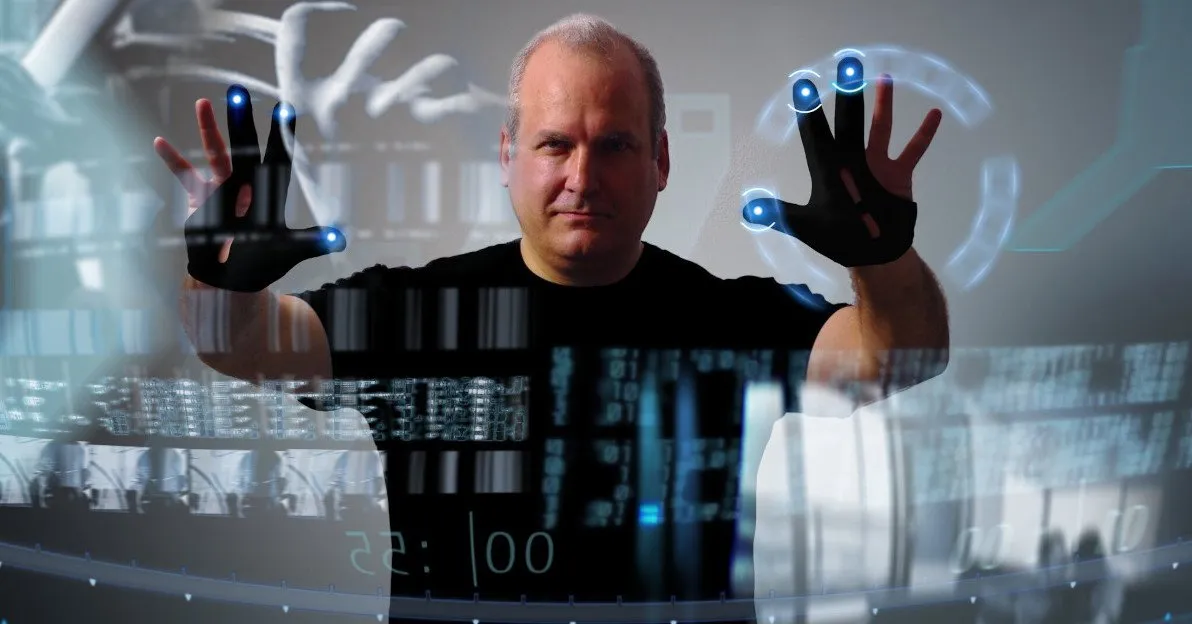 A futuristic, sci-fi scene: a man stands centered against a cool gray background, both hands raised as if pressing invisible controls. Black gloves with glowing blue points trace his fingertips, while translucent holographic arcs and data panels float around his hands. Soft reflections of code, grids, and interface elements layer across the image, making it feel like he’s physically manipulating a digital system in midair.